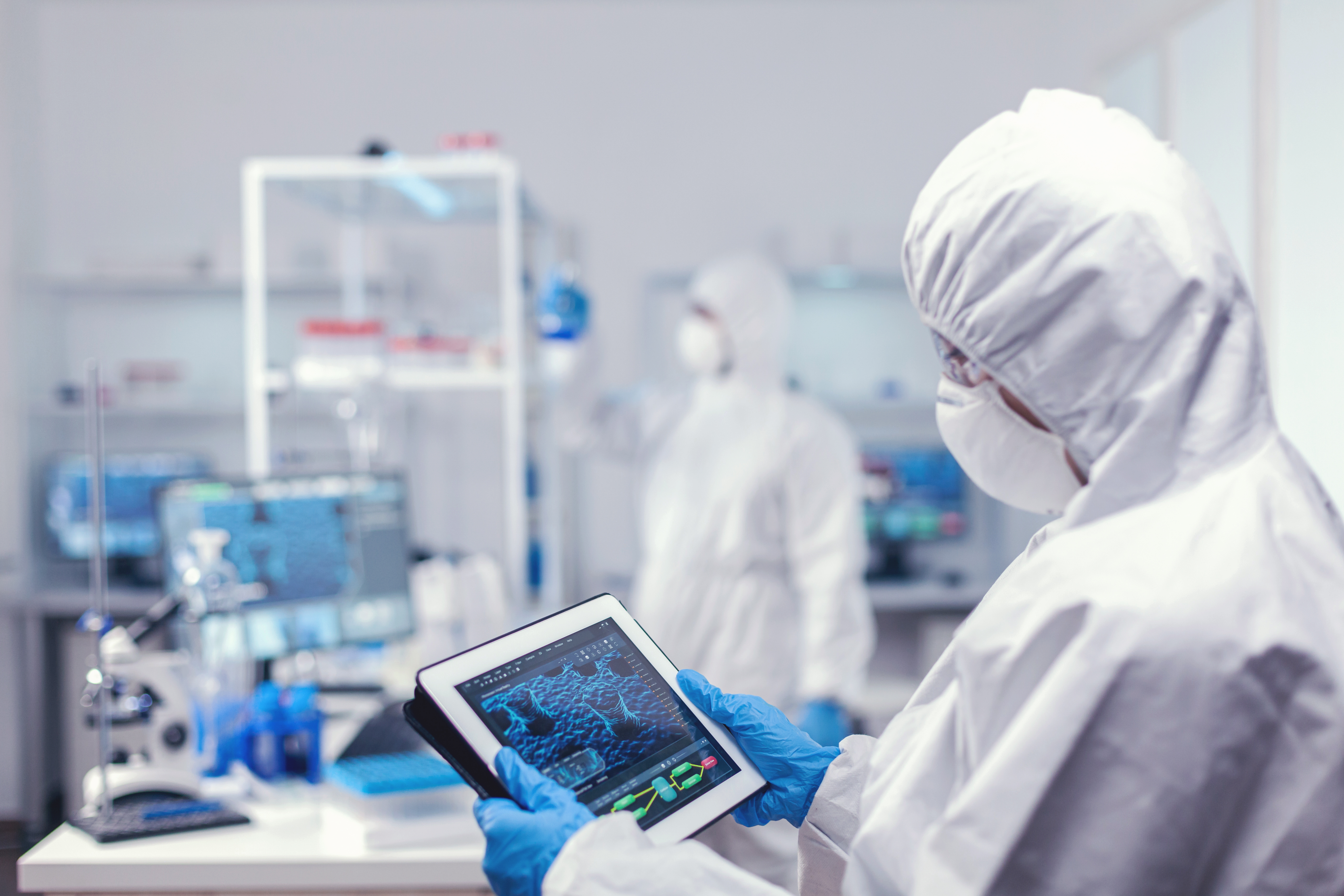 Concentrated medical researcher using digital tablet dressed in protective suit against infection with coronavirus. Team of scientists conducting vaccine development using high tech technology for researching treatment against covid19